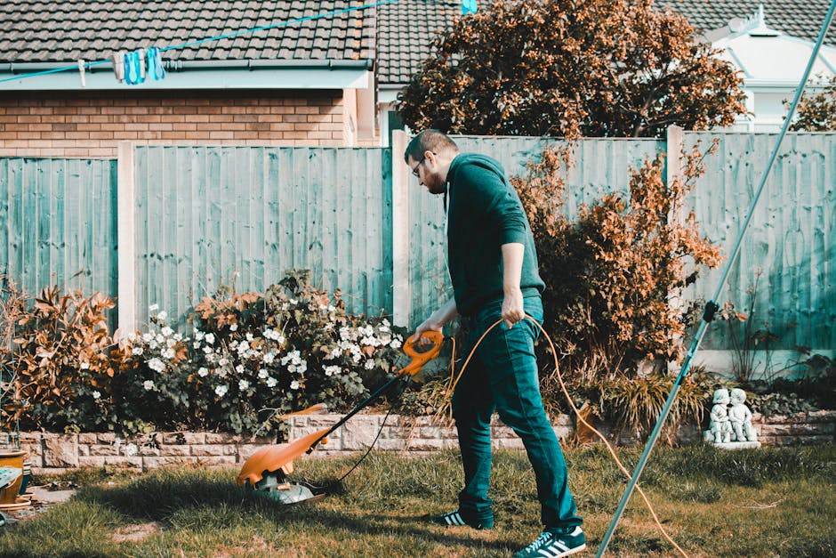 A man wearing glasses, a dark hoodie, and jeans is performing garden maintenance in a residential backyard. He is using an orange electric grass trimmer to cut the grass in a neatly kept lawn area, wi
